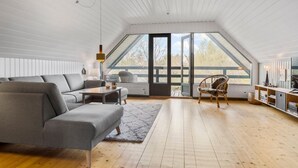 A spacious loft with a high ceiling, a large window, a grey sofa, a wooden floor, and a wicker chair. - 3 bedroom beautiful home in ørsted (Ørsted)