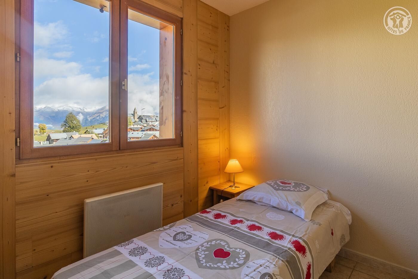 A cozy room with a bed, a window with a view of a town and mountains, and a lamp on a bedside table.