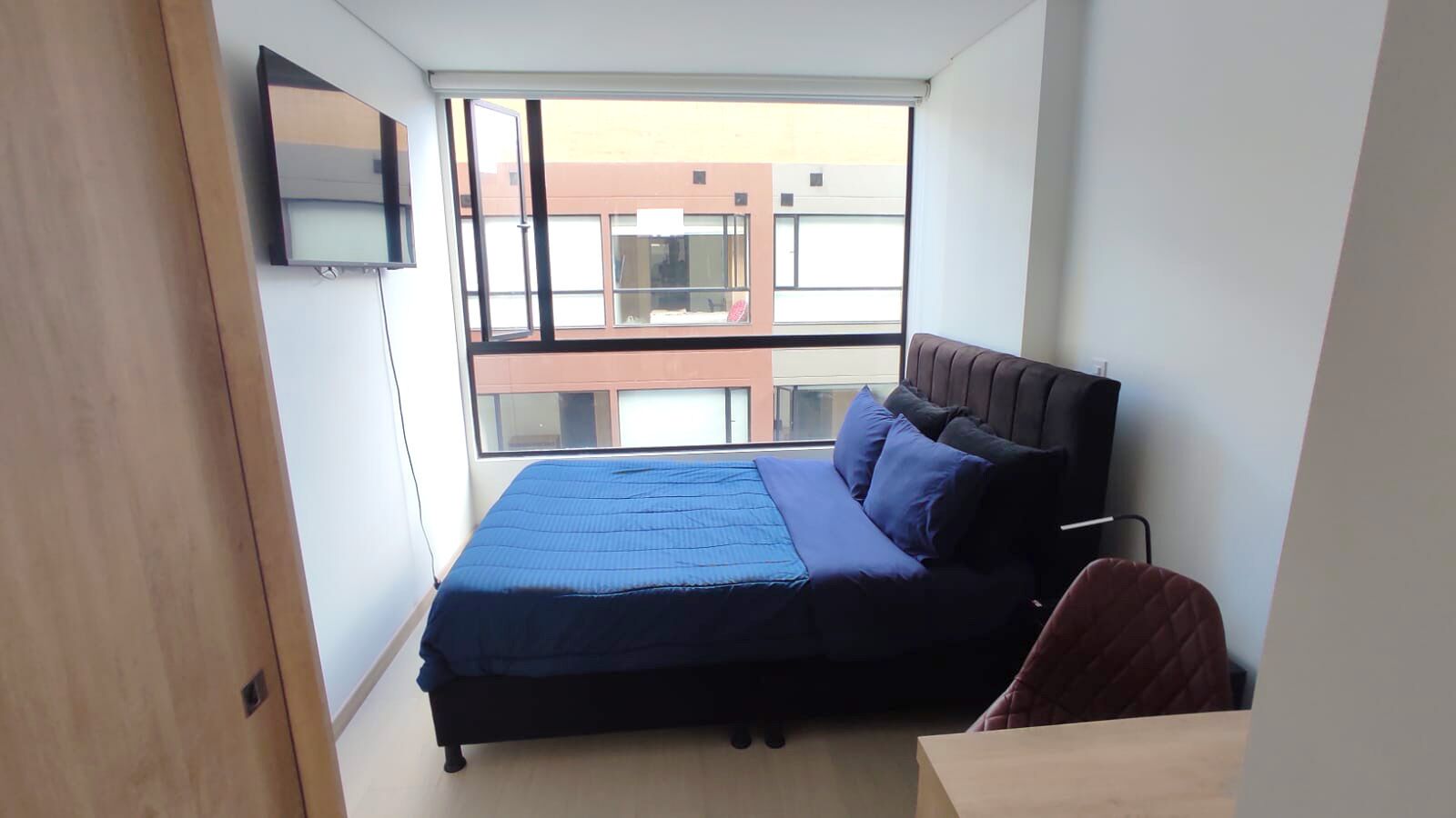 A bedroom with a bed, a TV mounted on the wall, and a window with a view of buildings.