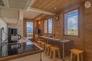 Une cuisine moderne équipée d’une machine à café, de tabourets en bois et de grandes fenêtres offrant une vue sur les montagnes.