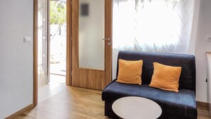 Una habitación con un sofá azul oscuro, dos cojines color mostaza, una mesita redonda y una puerta de madera que da al exterior.