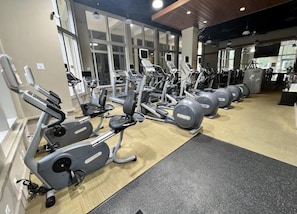 A gym with multiple treadmills and weight machines, large windows, and a wooden floor.