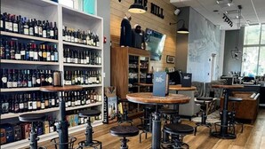 Zona de bar con suelos de madera, taburetes de metal y estanterías llenas de botellas de vino.