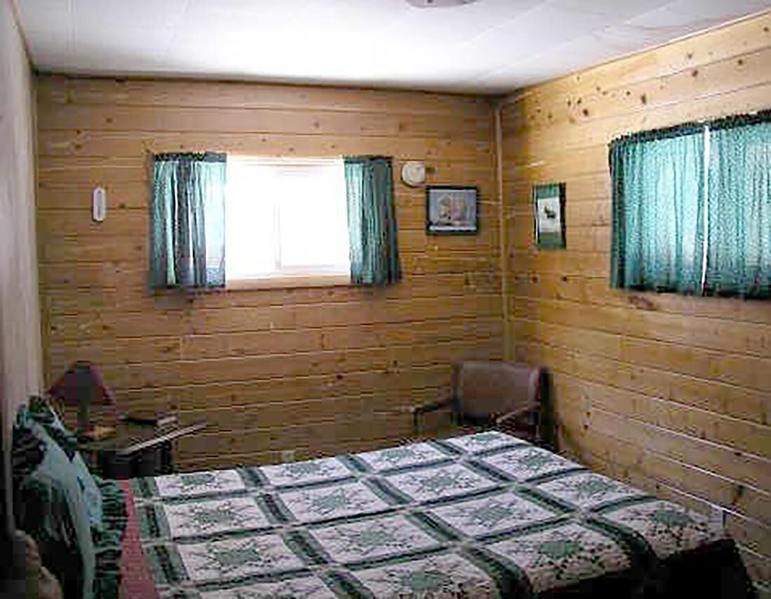 Ein Zimmer mit Holzwänden, einem Bett mit gemustertem Überwurf, einem Sessel, einem kleinen Tisch und zwei Fenstern mit Vorhängen.
