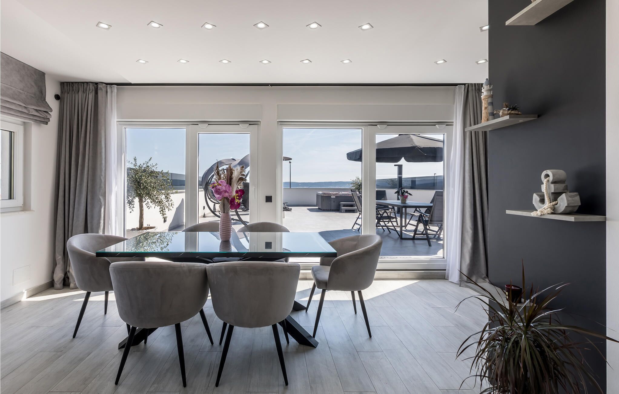 A modern dining area with a glass table and chairs, a view of the outdoors, and a ceiling with recessed lighting.
