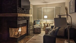 Un salon chaleureux avec une cheminée en pierre, un téléviseur à écran plat fixé au-dessus, un canapé confortable et une table d’appoint avec une lampe.