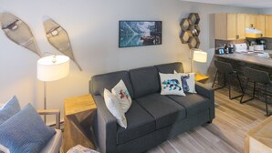 Un salon moderne avec un canapé gris foncé, deux tables d’appoint avec des lampes, une table basse en bois et un fauteuil bleu. On aperçoit à l’arrière-plan un coin cuisine avec des meubles en bois et un tabouret de bar.