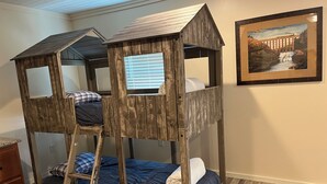 Una cama litera con estructura de madera y una escalera, un aparador y un cuadro en la pared.