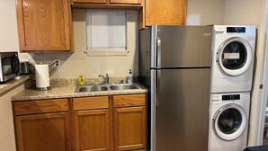 Una cocina con armarios de madera, un refrigerador de acero inoxidable, lavadora y secadora integradas, encimera de granito y una ventana con persianas.