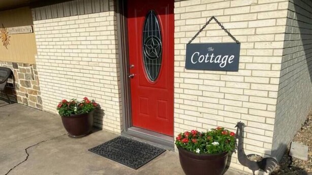 Una puerta roja con una reja de metal, un letrero que dice "The Cottage" y dos macetas con flores rojas en el porche.
