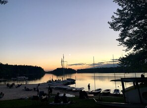 Une scène au bord d’un lac, avec des bateaux amarrés, des gens se détendant sur la rive et un ciel dégagé.