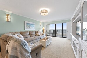A spacious living room with a large sofa, a coffee table, and a TV on a cabinet. There is a balcony with a view of the ocean. - Serenity by the Sea Direct Ocean Front Oasis w/Pool & Hot tub! (Cocoa Beach)