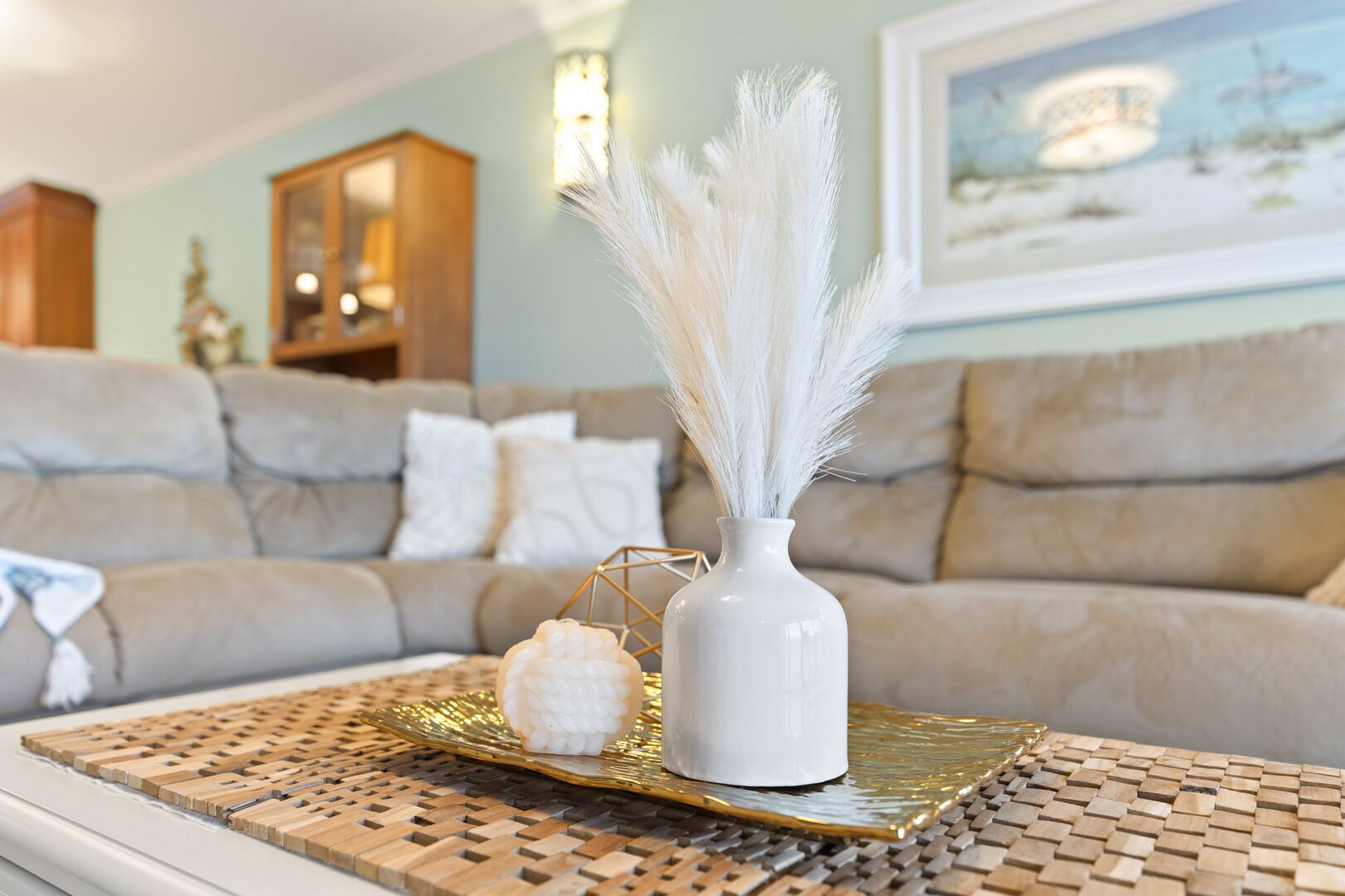 A living room with a beige sofa, a white vase with white feathers, and a decorative object on a woven tray.