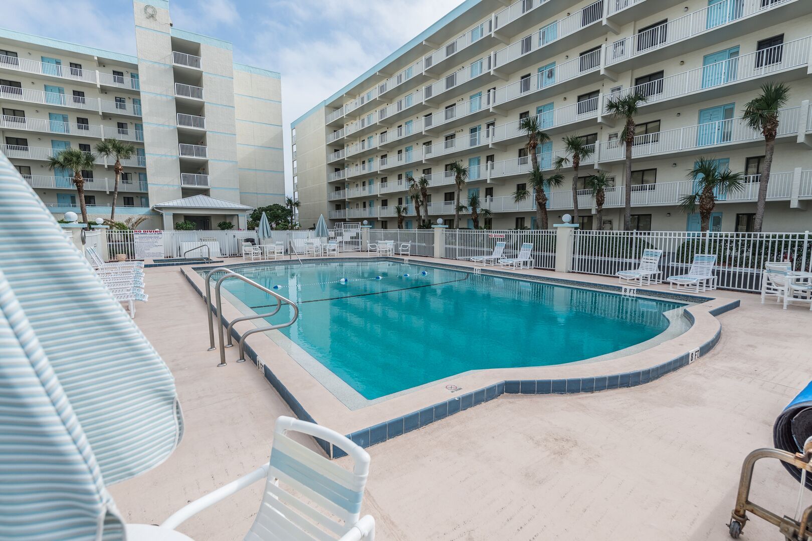 A hotel pool area with a rectangular pool, white lounge chairs, and a white fence.