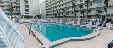 A hotel pool area with a rectangular pool, white lounge chairs, and a white fence.