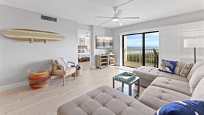A modern living room with a sofa, a chair, a small table, a ceiling fan, and a large window with a view of the ocean. - Seaside Escape | Modern 2BR w/ Balcony, Pool & Ocean Views!🌴 (Cape Canaveral)