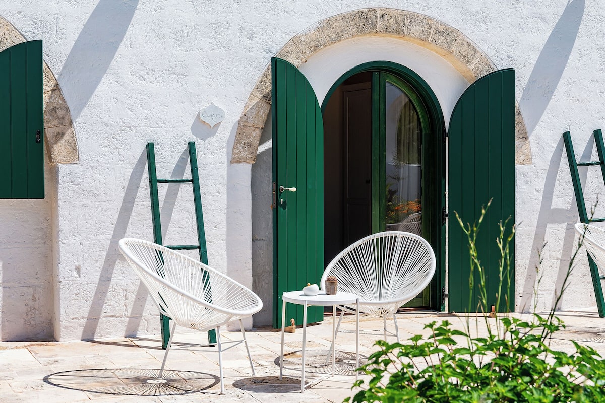 Un bâtiment blanc avec des portes et des fenêtres vertes, des chaises blanches et une petite table sur laquelle se trouve une théière.