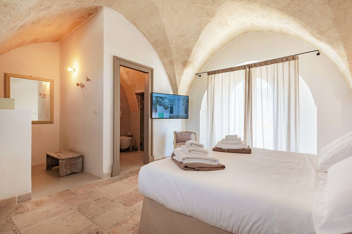 Une chambre à coucher avec un grand lit, une télévision, un petit tabouret en bois et une salle de bain visible à travers une arche.