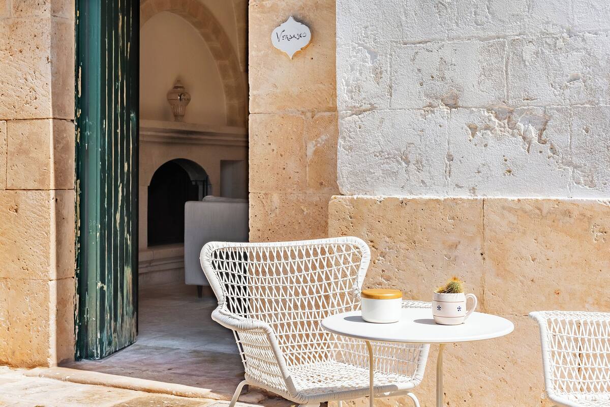 Un ensemble de chaise et de table en osier blanc est installé dans un espace extérieur ensoleillé, avec un mur de pierre et une porte ouverte donnant sur un espace intérieur.