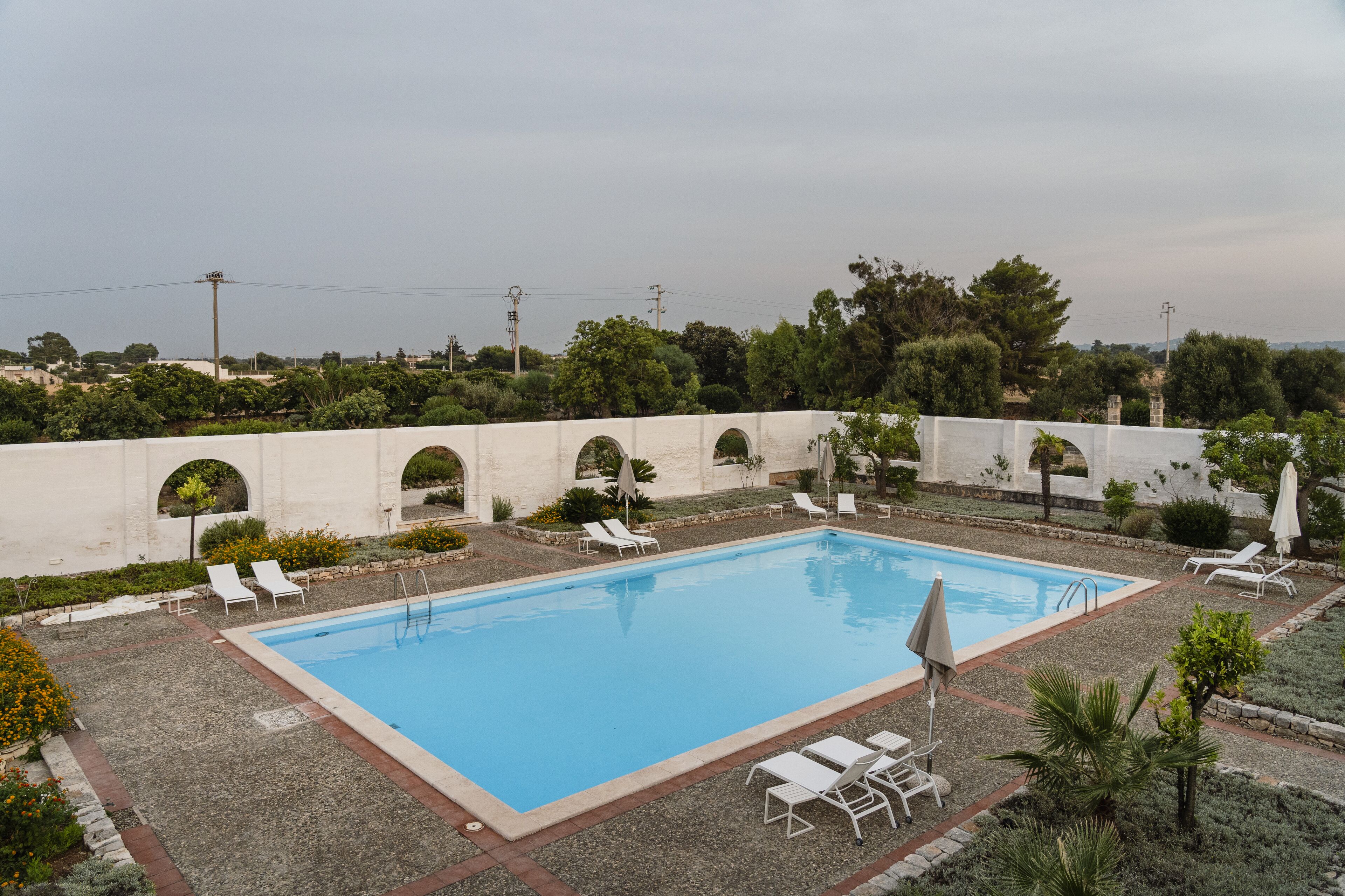 A rectangular swimming pool with white lounge chairs and umbrellas, surrounded by a low stone wall and greenery.