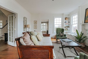 A spacious living room with a wooden sofa, a green armchair, and a coffee table. There is a bookshelf with books and decorative items, and a fireplace with a plant beside it. - The Cardinal House: Historic Home on Tree Streets (Waynesboro)