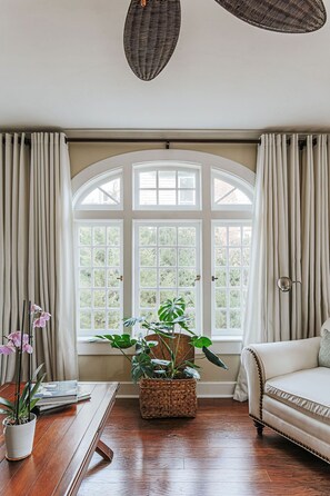 A room with a large arched window, a wooden floor, a white sofa, and a table with a plant and a vase. - The Cardinal House: Historic Home on Tree Streets (Waynesboro)
