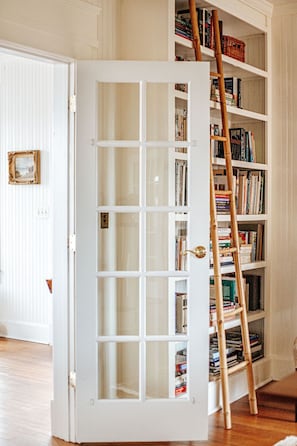 A white door with glass panels leading to a bookshelf filled with books. A wooden ladder leans against the bookshelf. - The Cardinal House: Historic Home on Tree Streets (Waynesboro)