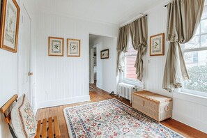 A room with a wooden bench, a patterned rug, a wicker chest, and framed botanical prints on the wall. - The Cardinal House: Historic Home on Tree Streets (Waynesboro)