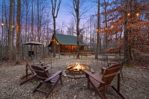 A cabin with a fire pit and outdoor seating, surrounded by trees and a clear sky.