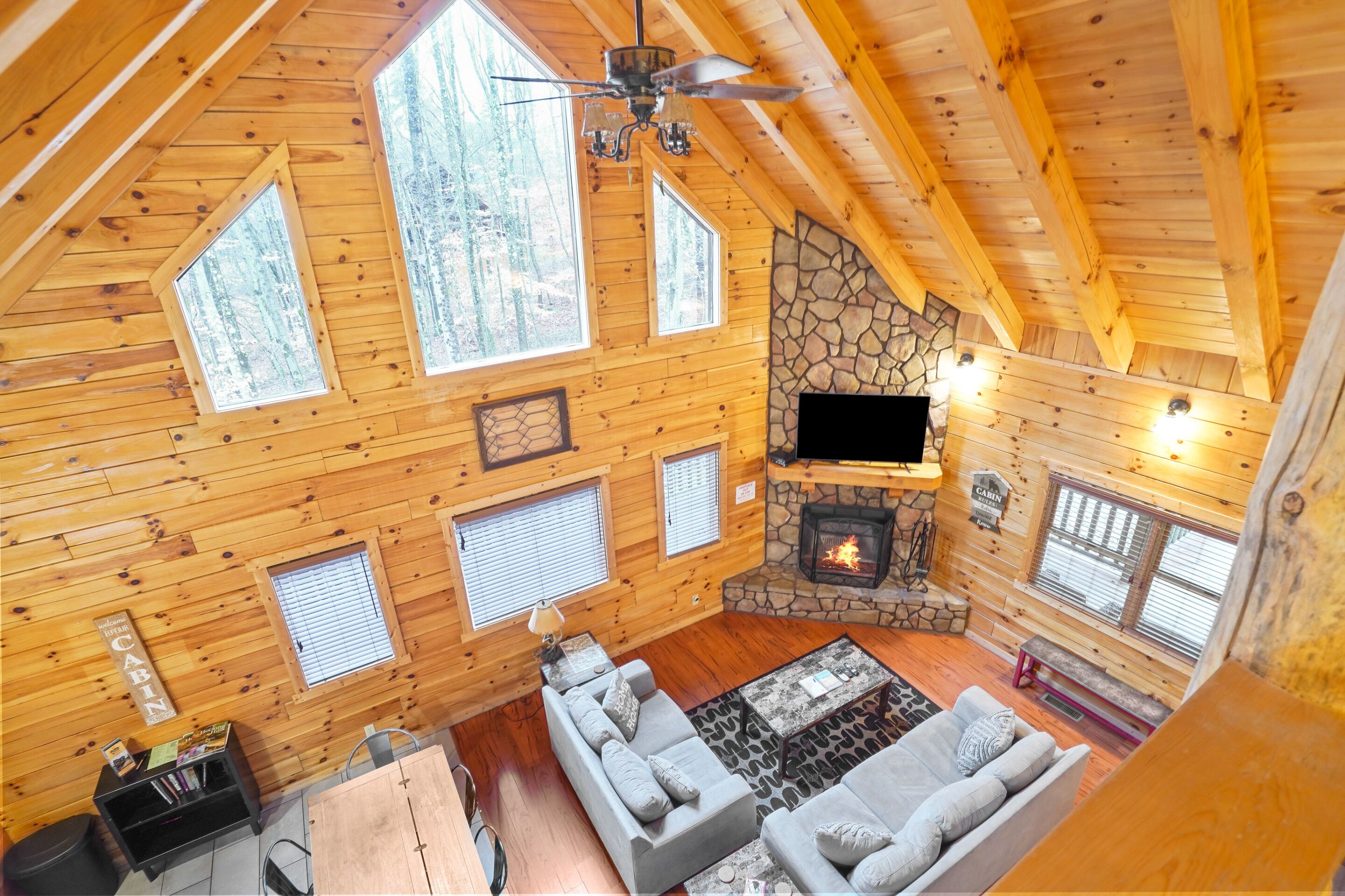A cabin interior with a stone fireplace, wooden walls, and a large window overlooking a forest.