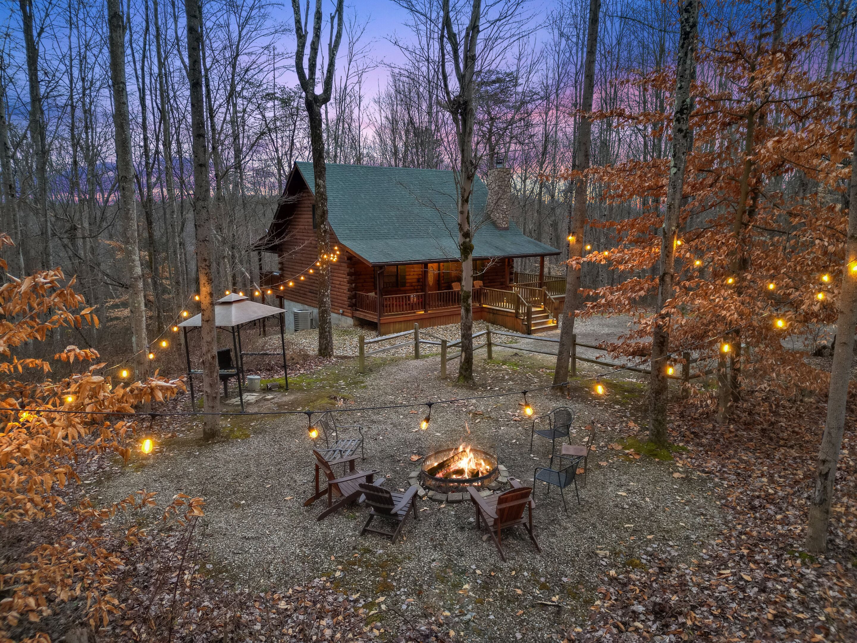 A cabin with a fire pit and outdoor seating, surrounded by trees and string lights.