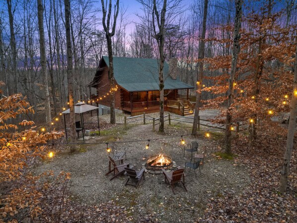 A cabin with a fire pit and outdoor seating, surrounded by trees and string lights.