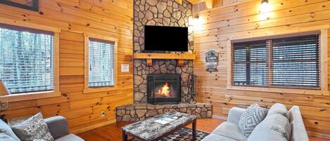 Interior de una cabaña con chimenea de piedra, paredes de madera, un sofá y un televisor.