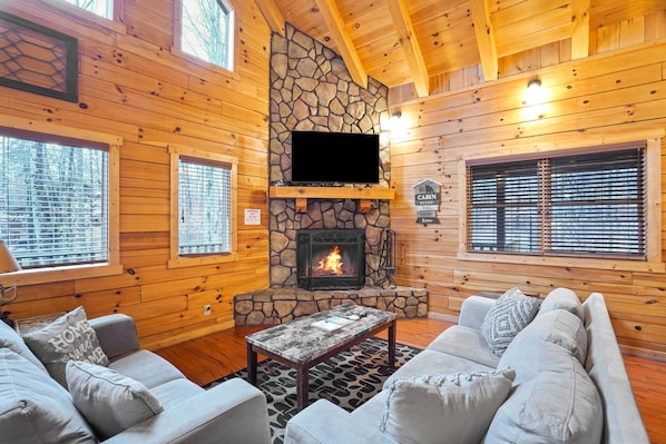 Interior de una cabaña con chimenea de piedra, paredes de madera, un sofá y un televisor.