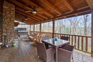 A covered deck with a stone fireplace, wicker furniture, and a view of the surrounding forest.