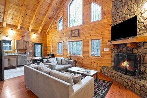 A cozy log cabin interior with a stone fireplace, wooden walls, a sofa, and a dining area. - Fireside Cabin | Hocking Hills, Hot Tub, Sleeps 8 (Rockbridge)
