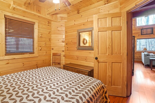 A wooden cabin room with a bed, a chair, a small table, and a door leading to another room.