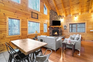 A cabin interior with a stone fireplace, wooden walls, a TV mounted above it, a dining table with chairs, and a sofa.