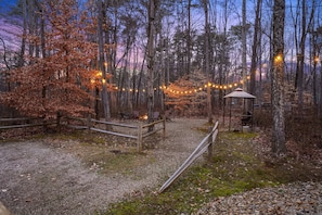 A forested area with a wooden deck, a fire pit, and string lights.