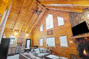 A cozy cabin with wooden walls, a stone fireplace, a ceiling fan, and a kitchen area.