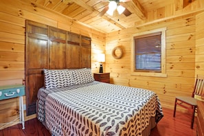 A wooden cabin bedroom with a bed, a nightstand, a lamp, a window with blinds, and a wooden wall with a circular decoration.