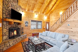 A cozy cabin interior with a stone fireplace, a flat-screen TV mounted above it, a gray sofa, and a wooden staircase.