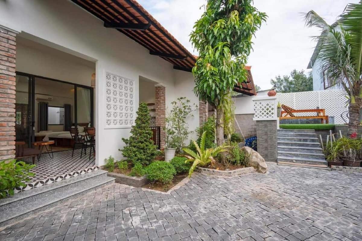 A house with a tiled patio, a covered outdoor seating area, and a garden with various plants and a palm tree.
