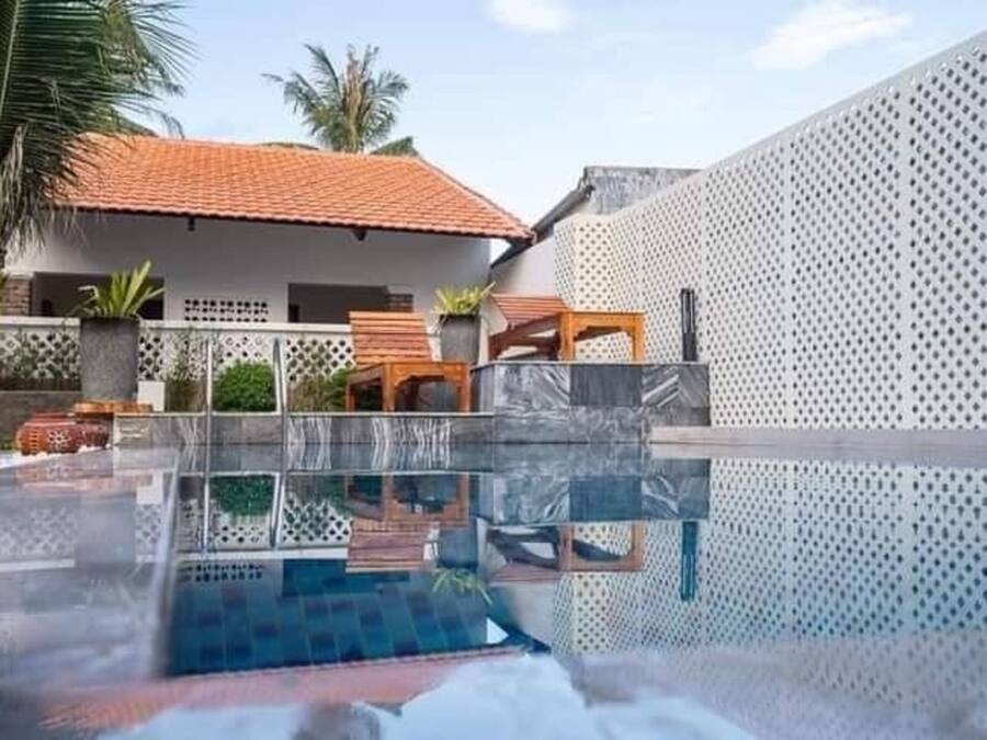 A swimming pool with a clear view of the surrounding area, including a house with a tiled roof and a white wall with a perforated pattern.