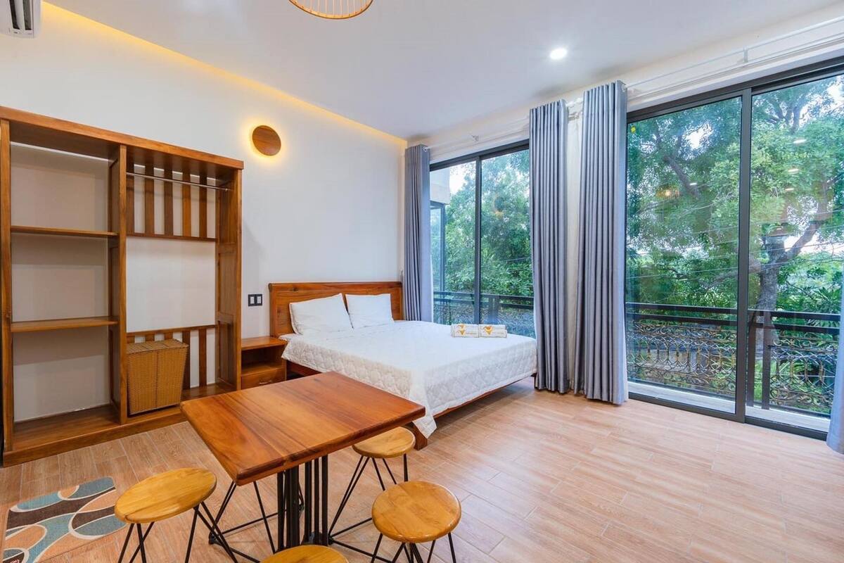 Un dormitorio con una cama, una mesa de madera con taburetes, un armario y un amplio ventanal con cortinas.