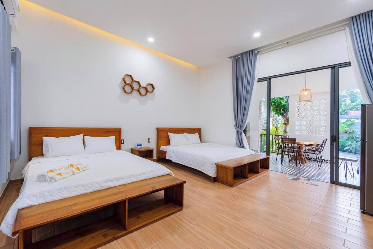 Habitación de hotel con dos camas, suelo de madera, balcón con mesa y sillas y vistas a zonas verdes.