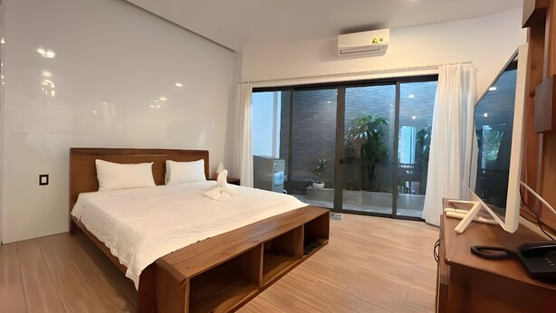 A modern bedroom with a wooden bed, a built-in bench, a large sliding glass door leading to a balcony, and a ceiling-mounted air conditioning unit. - Nhu House Apartment Phu Quoc  (Phu Quoc)