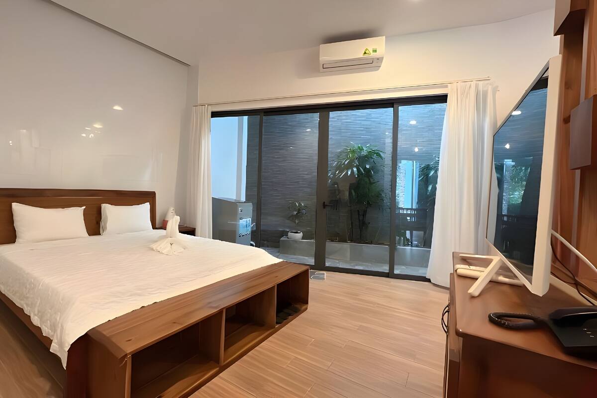 Un dormitorio moderno con una cama de madera, un banco empotrado, un amplio ventanal corredero que da a un balcón y un equipo de aire acondicionado instalado en el techo.
