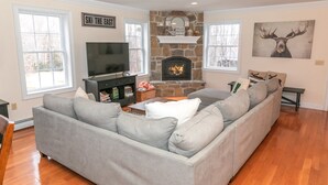 A living room with a stone fireplace, a gray sofa, a flat-screen TV, and a sign that reads "SKI THE BEST". - Spacious Chalet - PetFriendly/HotTub/Firepit/GameRoom/AC (Wilmington)