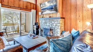 A cozy living room with a stone fireplace, wooden walls, and a large window overlooking a deck.